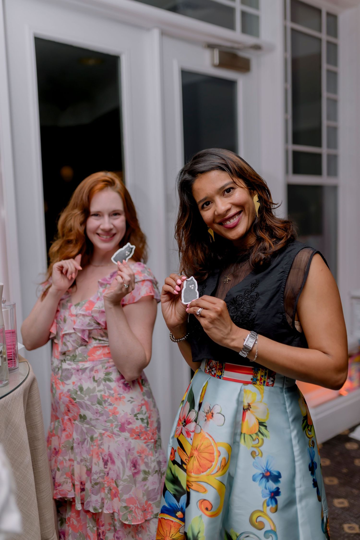 Two women in floral dresses smile and hold small cards at an elegant indoor event.