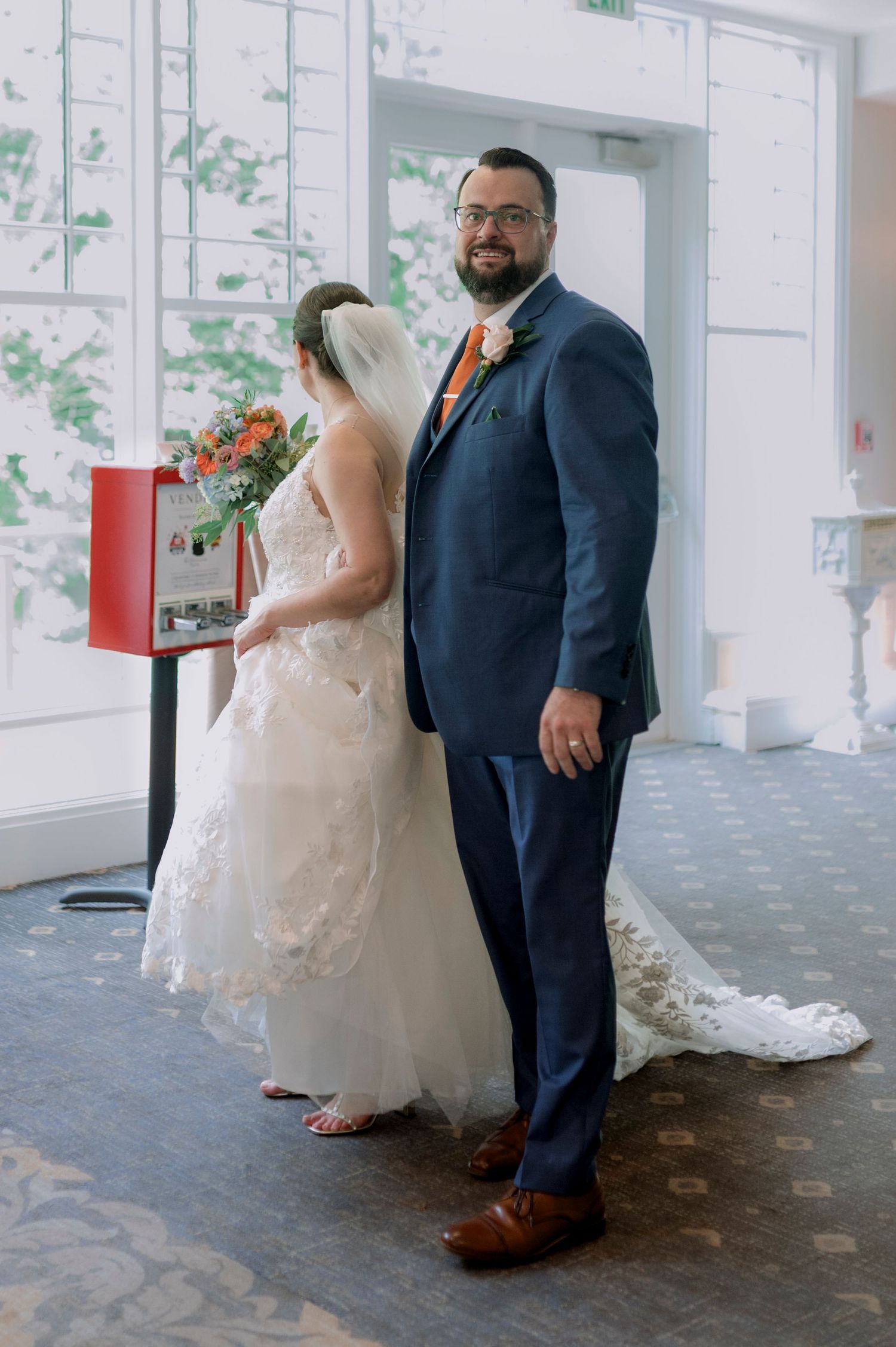 Groom in navy suit with orange boutonniere stands beside bride in lace wedding gown holding floral bouquet indoors.