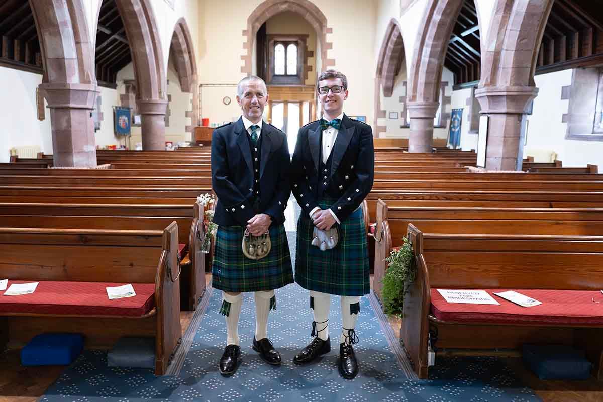 Two men in traditional Scottish kilts standing together in a historic stone church with wooden pews.