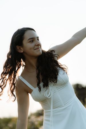 Young woman in white dress smiling with arm raised outdoors at golden hour.