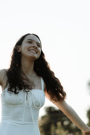 Young woman with long dark hair smiling joyfully in a white dress outdoors against a bright sky.