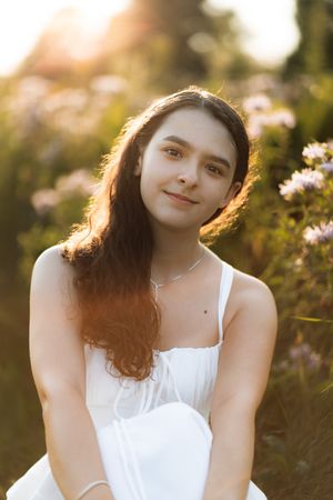 Young woman in white dress sitting in a sunlit wildflower field, smiling softly with golden hour light.