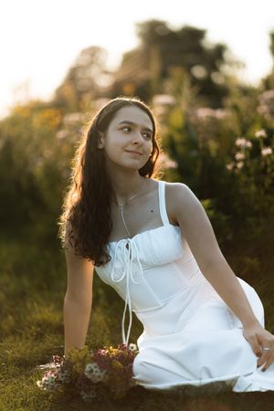 Young woman in white dress sitting in a sunlit meadow with wildflowers, smiling softly in golden hour light.