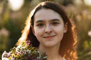 Young woman with dark hair smiling softly, holding a wildflower bouquet in a sunlit field.