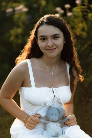 Young woman in white dress holding a blue stuffed bunny, smiling outdoors in soft golden hour light.