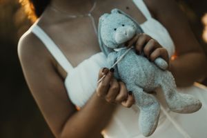 A person in a white dress holds a soft blue stuffed bunny toy with a bow, in a warm, softly lit setting.