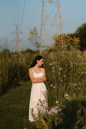Woman in white dress standing among wildflowers in a field with power lines in the background at golden hour.