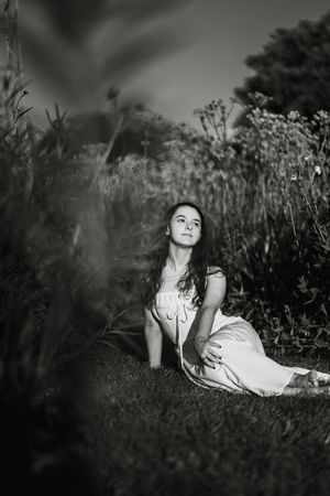 Black and white photo of a young woman in a white dress sitting gracefully in a lush garden meadow.
