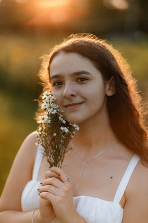 Young woman holding white daisies in golden sunset light, wearing a white dress and delicate necklace.