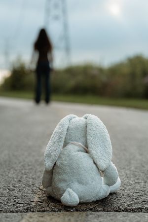 Abandoned white stuffed bunny toy on road with blurred girl walking away in background.
