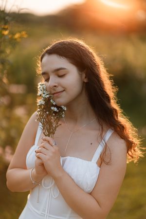 Young girl in white dress smelling wildflowers at golden sunset in a meadow.