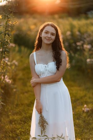 Young woman in white sundress holding wildflowers in golden sunset light in a field.