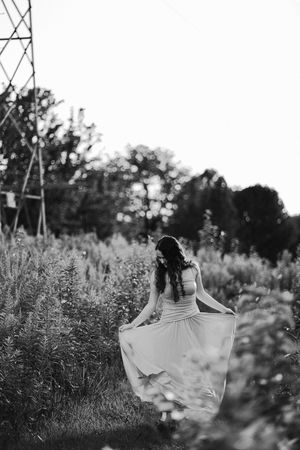Black and white photo of a woman in a flowing dress walking through a wildflower field near a metal tower.