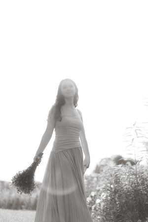 Black and white photo of a woman in a flowing dress holding wildflowers, standing outdoors in soft light.