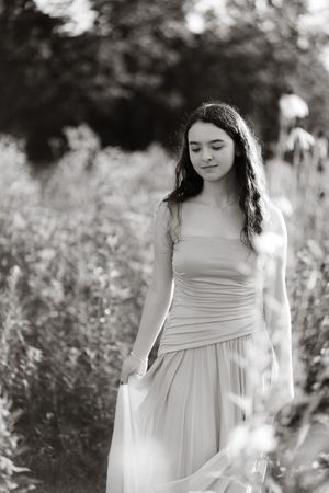 Black and white portrait of a young woman in a flowing dress standing among wildflowers in a garden.