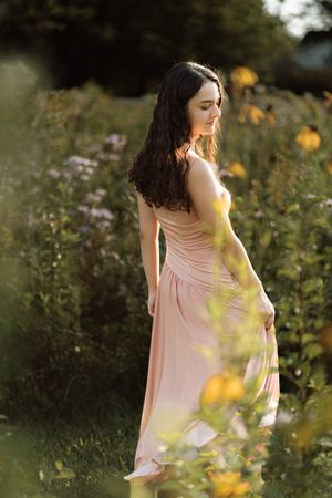 Young woman in a flowing pink dress standing among wildflowers in a garden during golden hour.