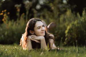 Young woman lying on grass in a sunlit park, resting her chin on her hands, gazing thoughtfully into the distance.