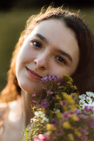 Young woman with brown hair smiling softly while holding a bouquet of wildflowers in warm sunlight.