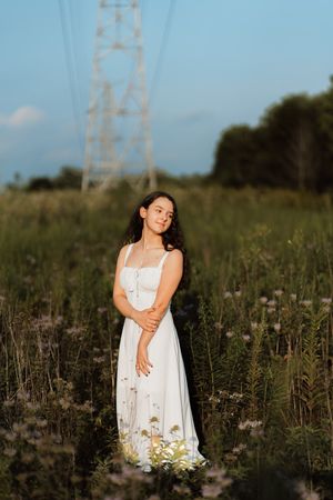 Young woman in white dress standing in wildflower field at golden hour with power tower in background.