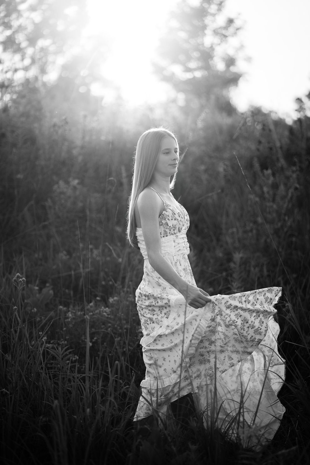 Black and white photo of a young woman in a floral dress standing in a sunlit field.