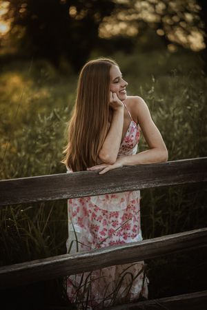 Young woman in floral dress leaning on wooden fence, smiling at golden hour sunset with lush green background.