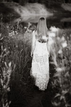 Black and white photo of a woman in a floral maxi dress walking through a wildflower meadow toward a serene lake.