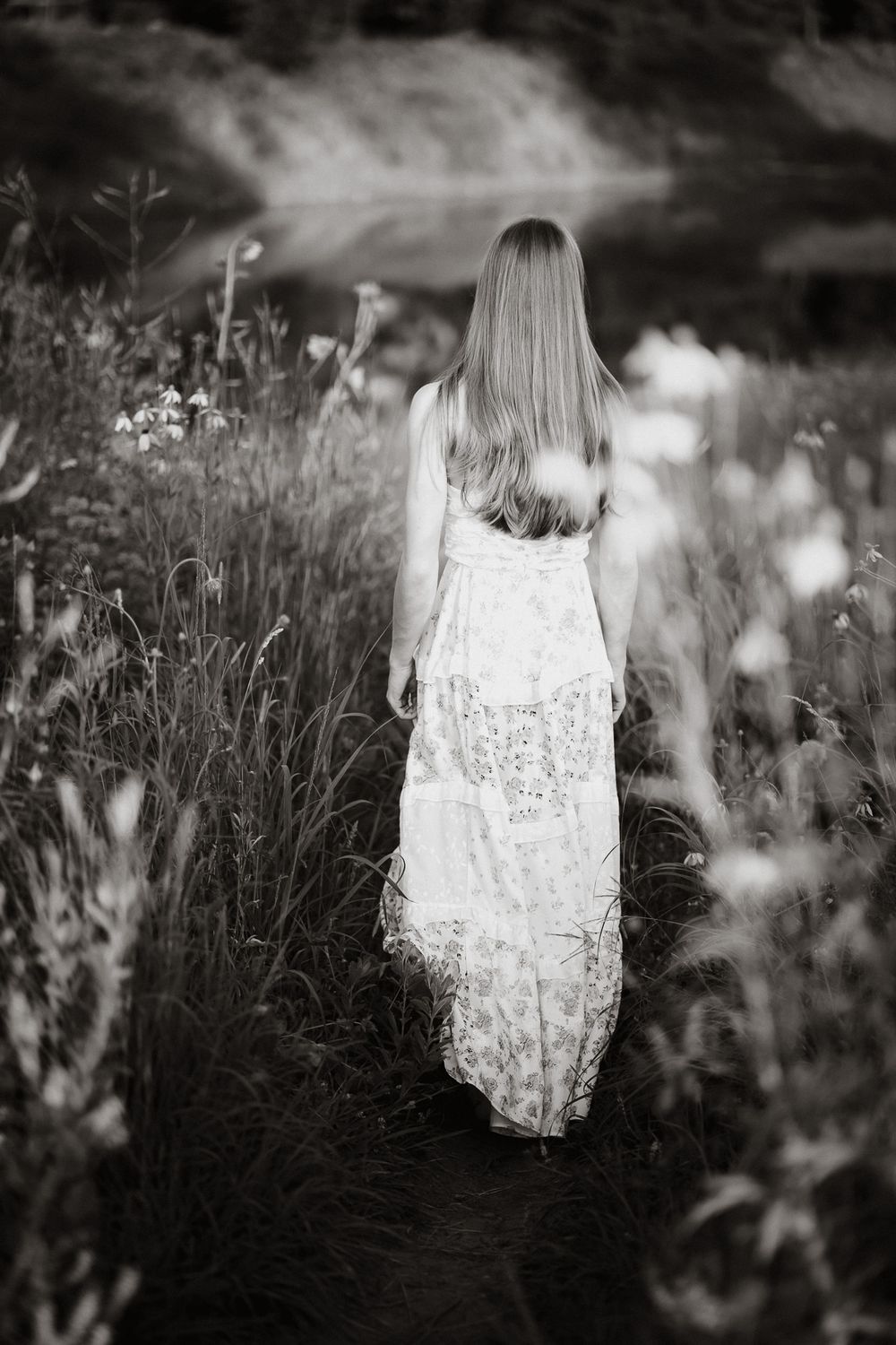 Black and white photo of a woman in a floral maxi dress walking through a wildflower meadow toward a serene lake.