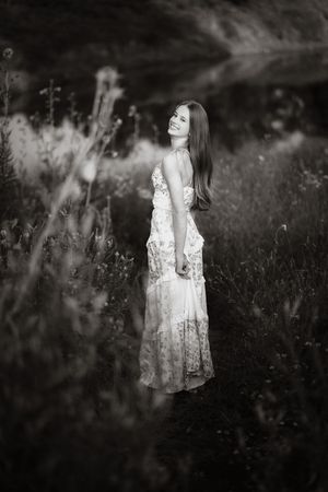 Black and white photo of a smiling woman in a floral maxi dress standing among wildflowers in a meadow.