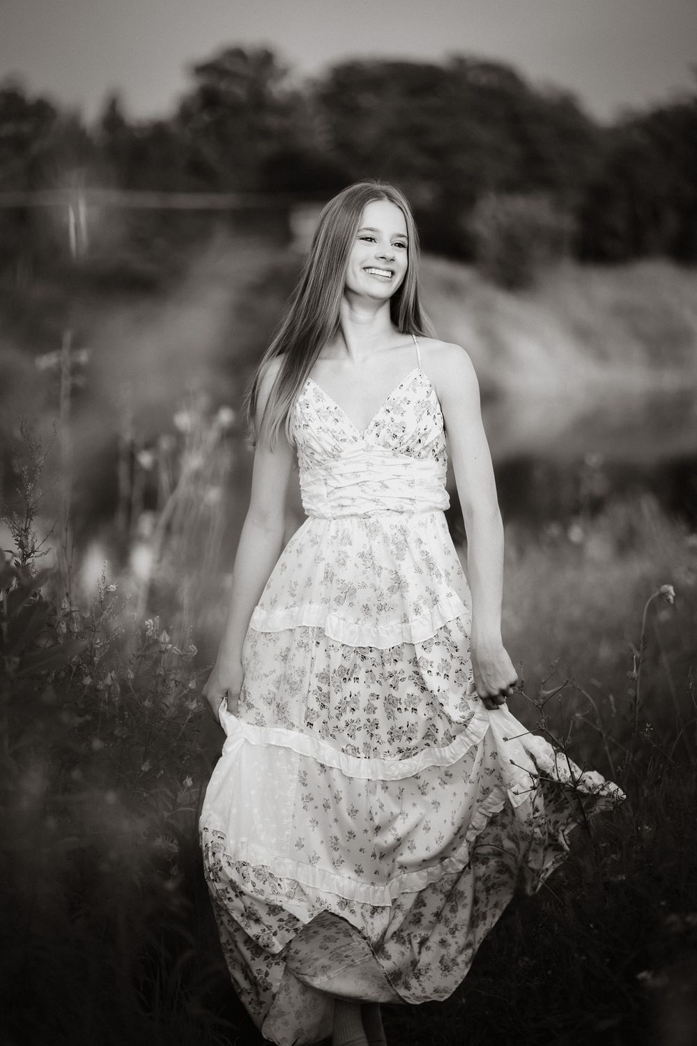 Black and white photo of a smiling young woman in a floral dress standing in a field of wildflowers.