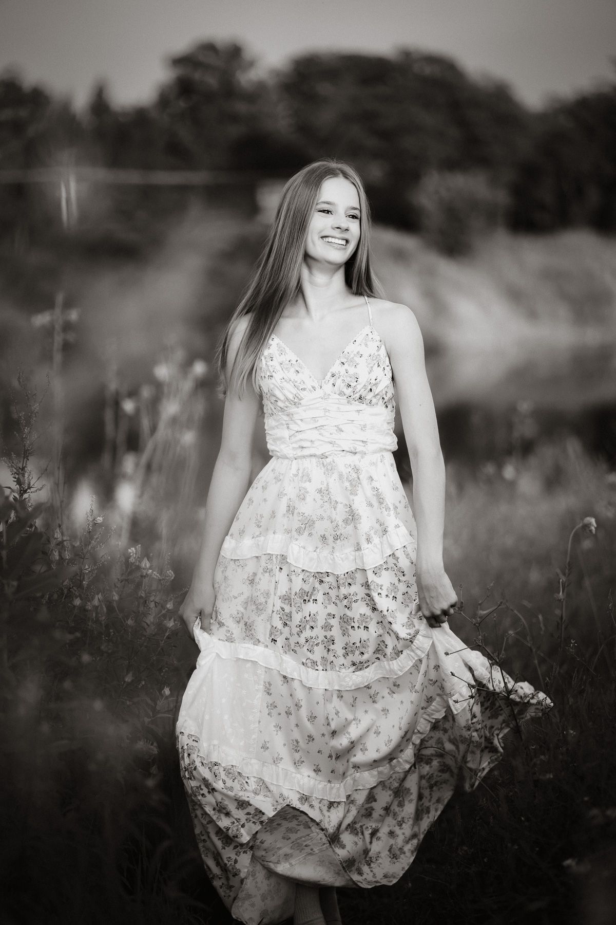 Black and white photo of a smiling young woman in a floral dress standing in a field of wildflowers.