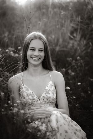 Black and white portrait of a smiling young woman sitting in a field wearing a floral dress.
