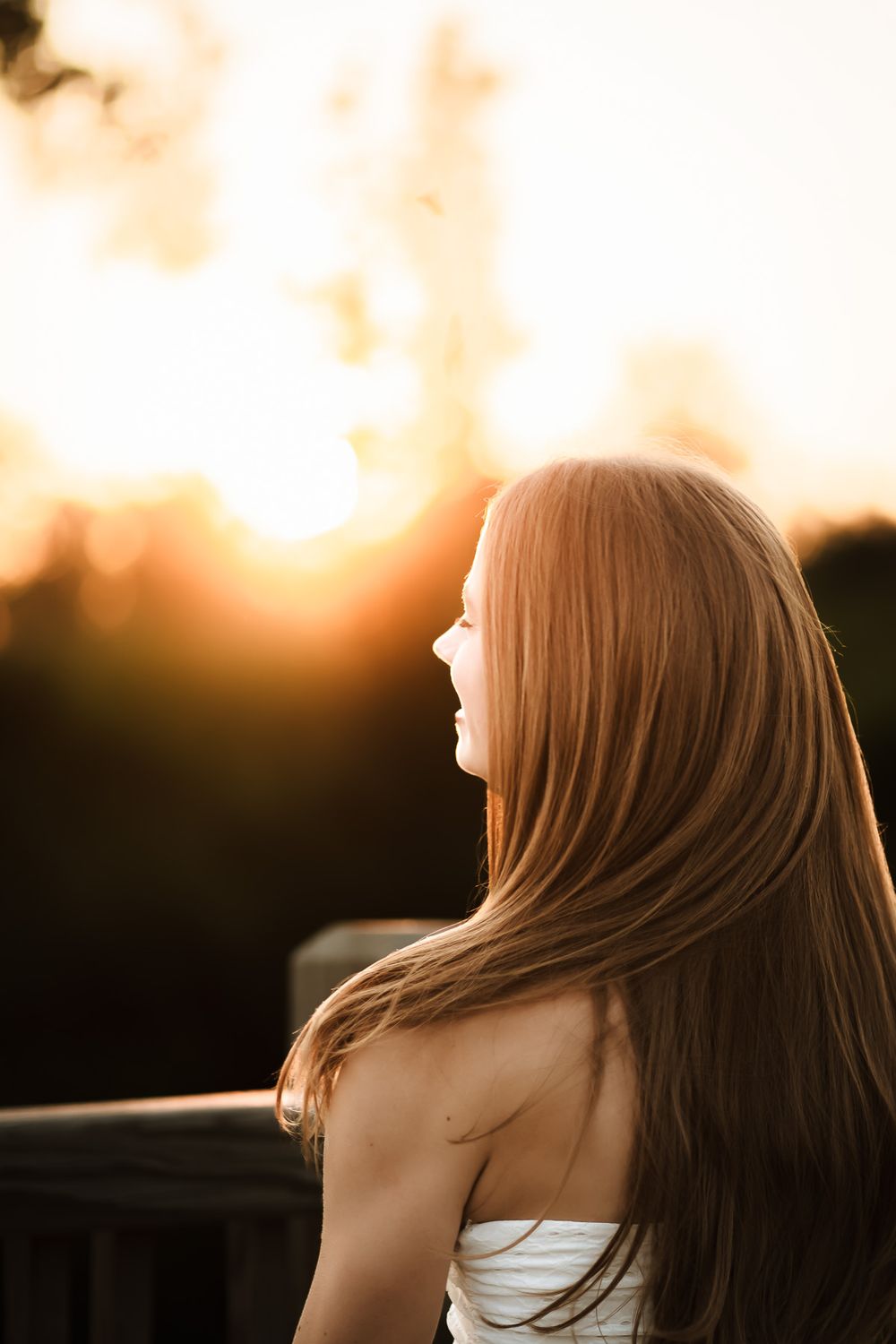 Young woman with long auburn hair gazes at golden sunset from outdoor deck, backlit by warm glowing light.