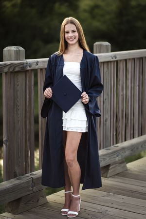 Smiling teenage girl in graduation cap and gown holding diploma on wooden bridge outdoors.