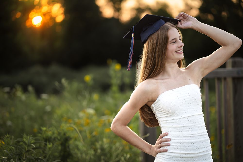 Young woman in white dress holding graduation cap outdoors at sunset, celebrating academic achievement.
