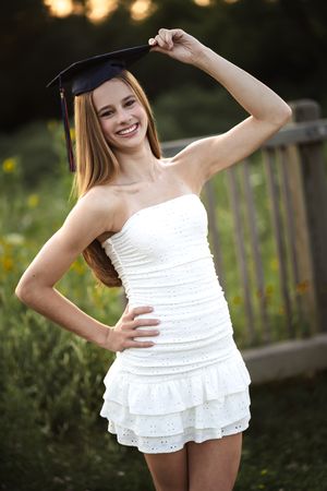 Smiling young woman in white strapless dress holding graduation cap outdoors in golden hour light.