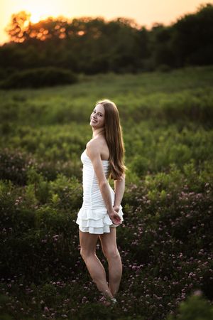Young woman in white ruffled mini dress posing in flower field at golden sunset.