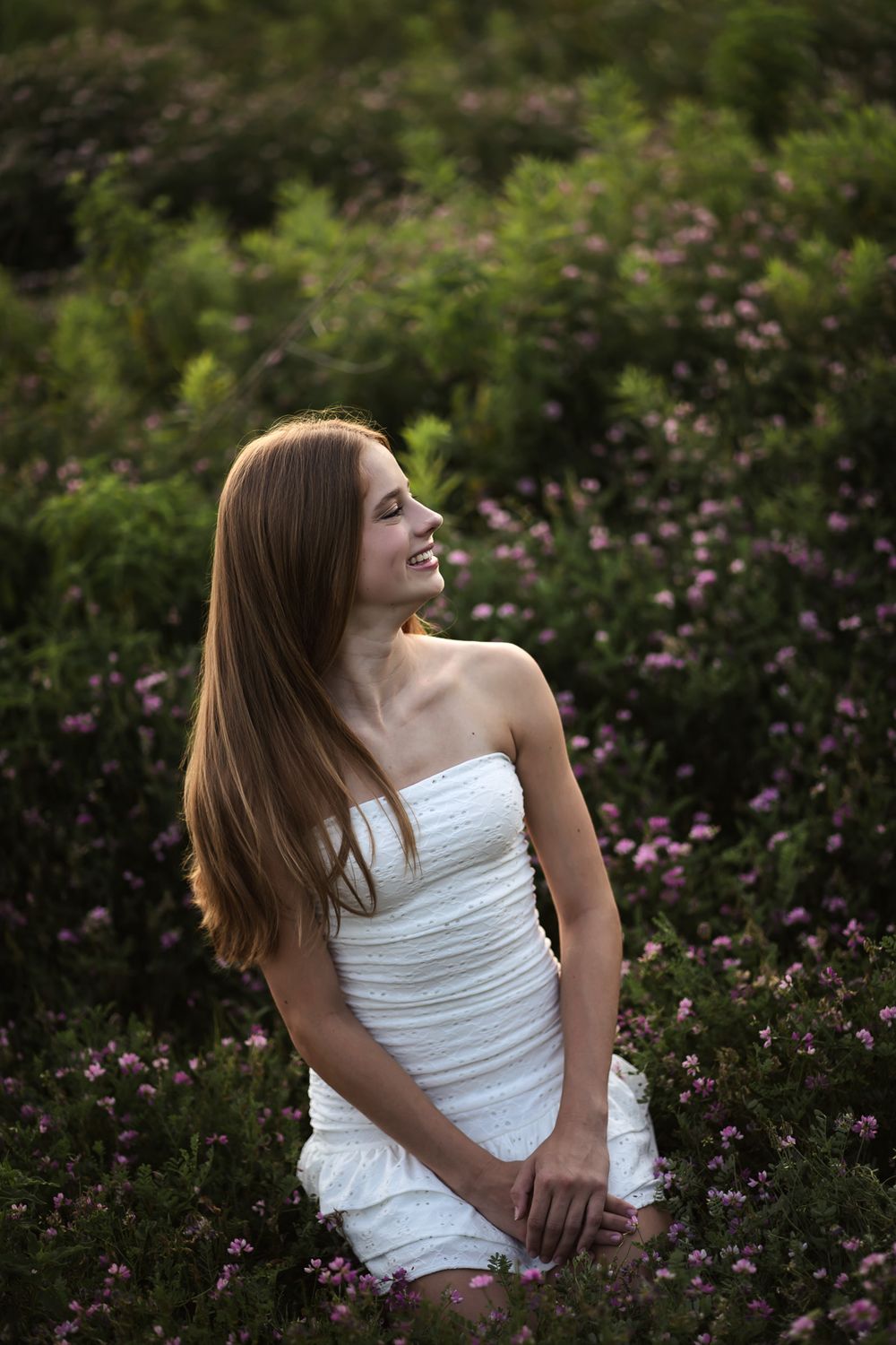 Young woman in white strapless dress smiling among pink wildflowers and green bushes outdoors.