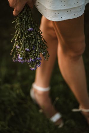 Woman in white dress holding a bouquet of purple wildflowers while standing on green grass wearing white heeled sandals.