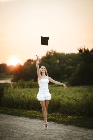 Ballerina in white dress tossing graduation cap at sunset outdoors.