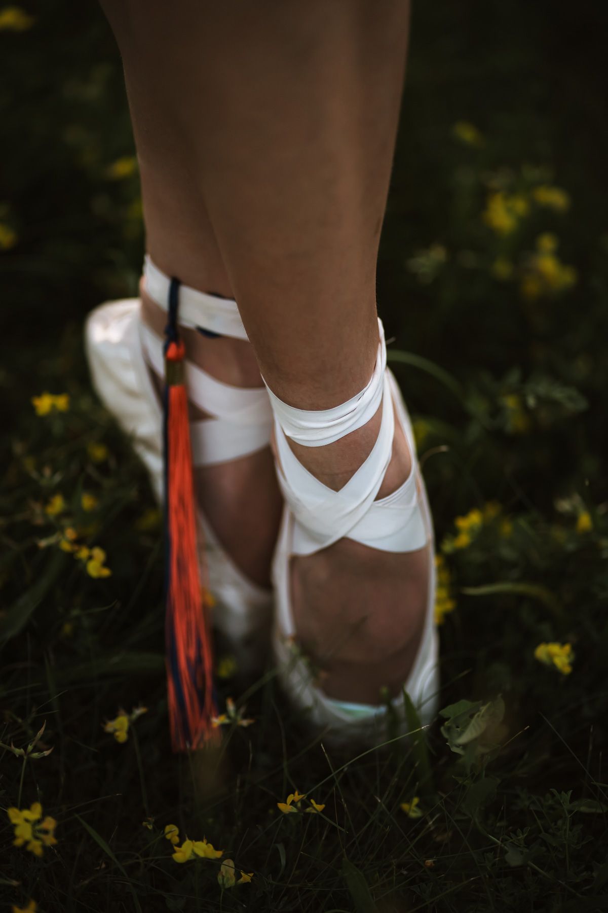 Close-up of ballet pointe shoes wrapped in white ribbons with colorful tassels, standing on green grass with yellow flowers.