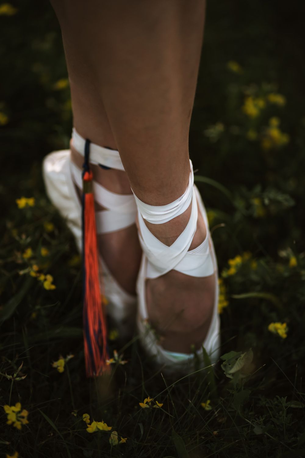 Close-up of ballet pointe shoes wrapped in white ribbons with colorful tassels, standing on green grass with yellow flowers.