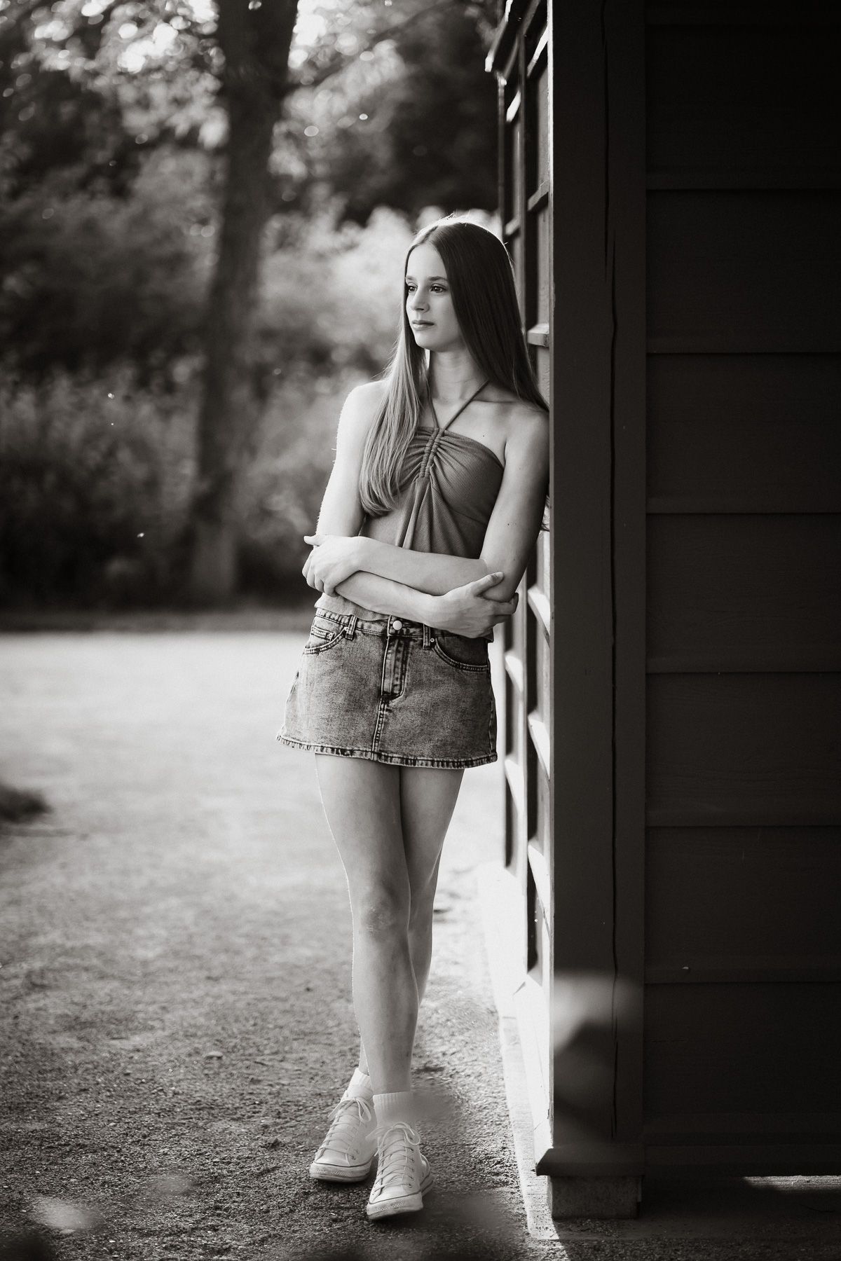 Black and white portrait of a young woman in denim skirt leaning against a building outdoors.