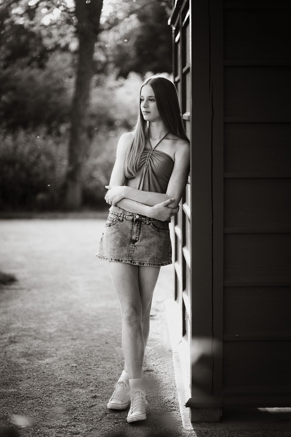 Black and white portrait of a young woman in denim skirt leaning against a building outdoors.