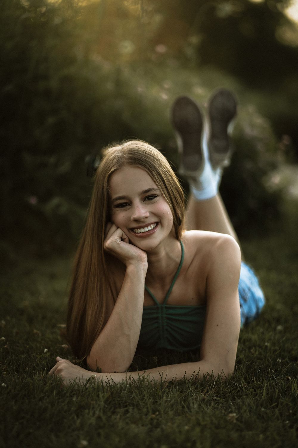 Smiling young woman with long blonde hair lying on green grass in a blue dress and sneakers, bathed in warm sunlight.