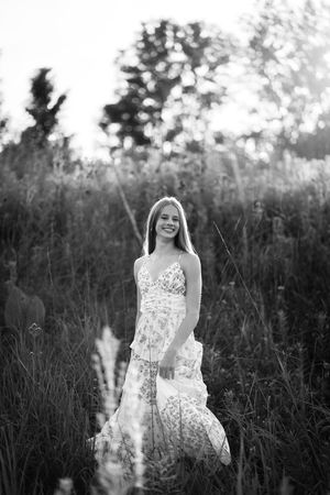 Young woman in floral maxi dress smiling in tall grass field, black and white portrait photography.