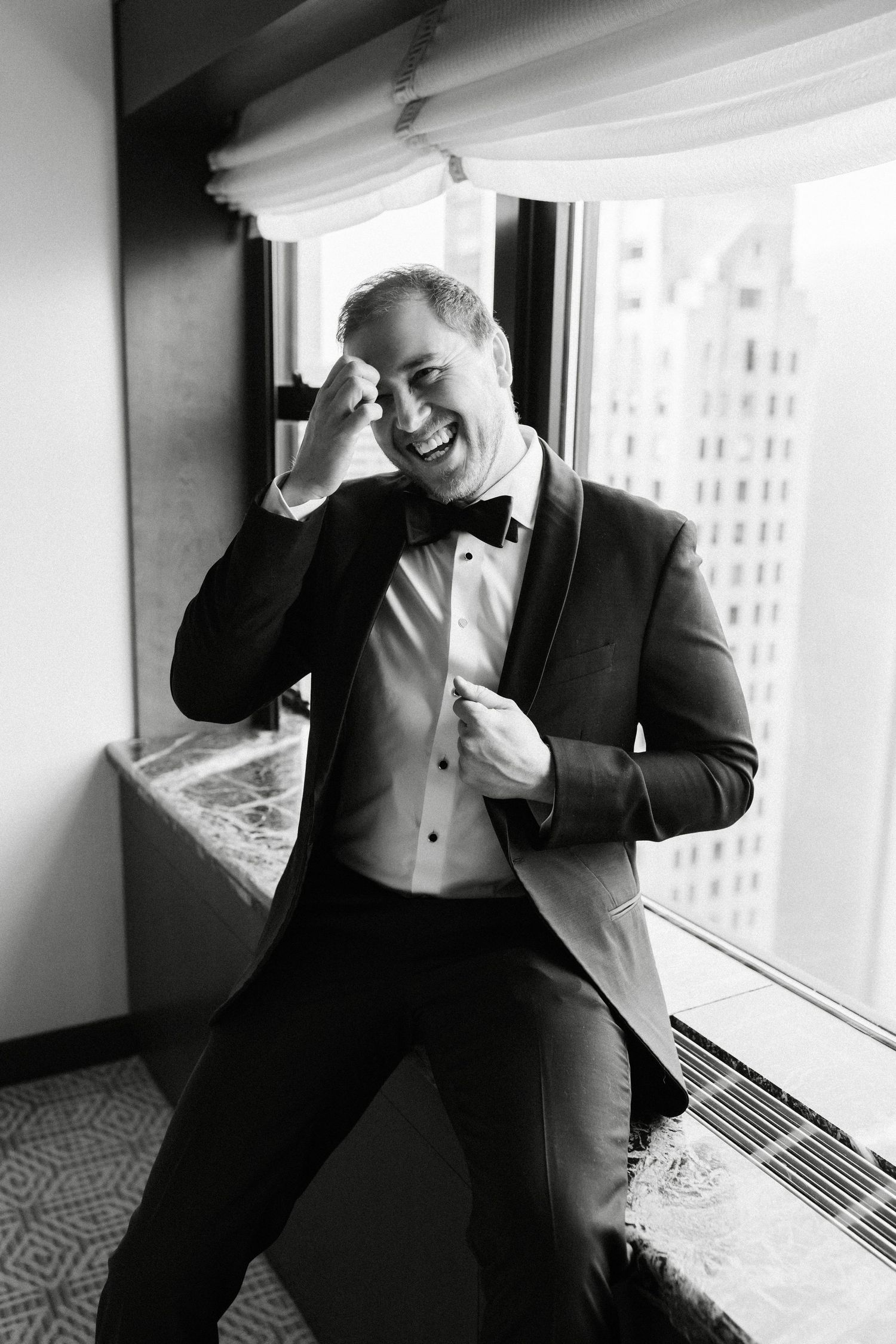 Joyful groom in black tuxedo and bow tie laughing by hotel window in black and white wedding photo.