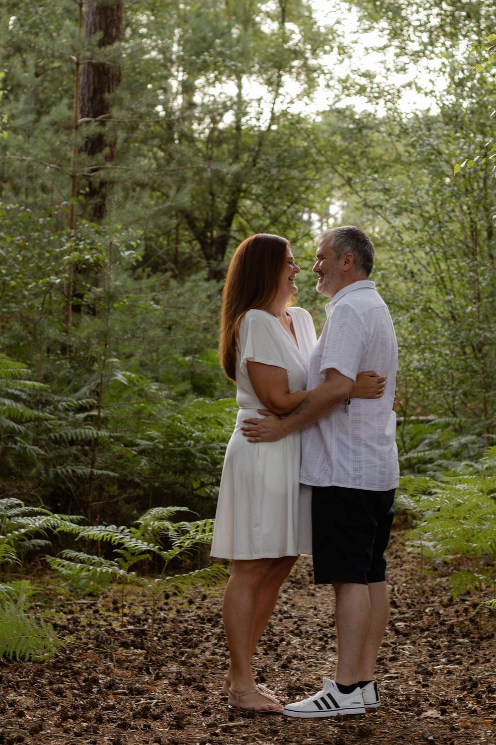 A couple in white outfits embrace on a forest path surrounded by lush green ferns and tall trees.