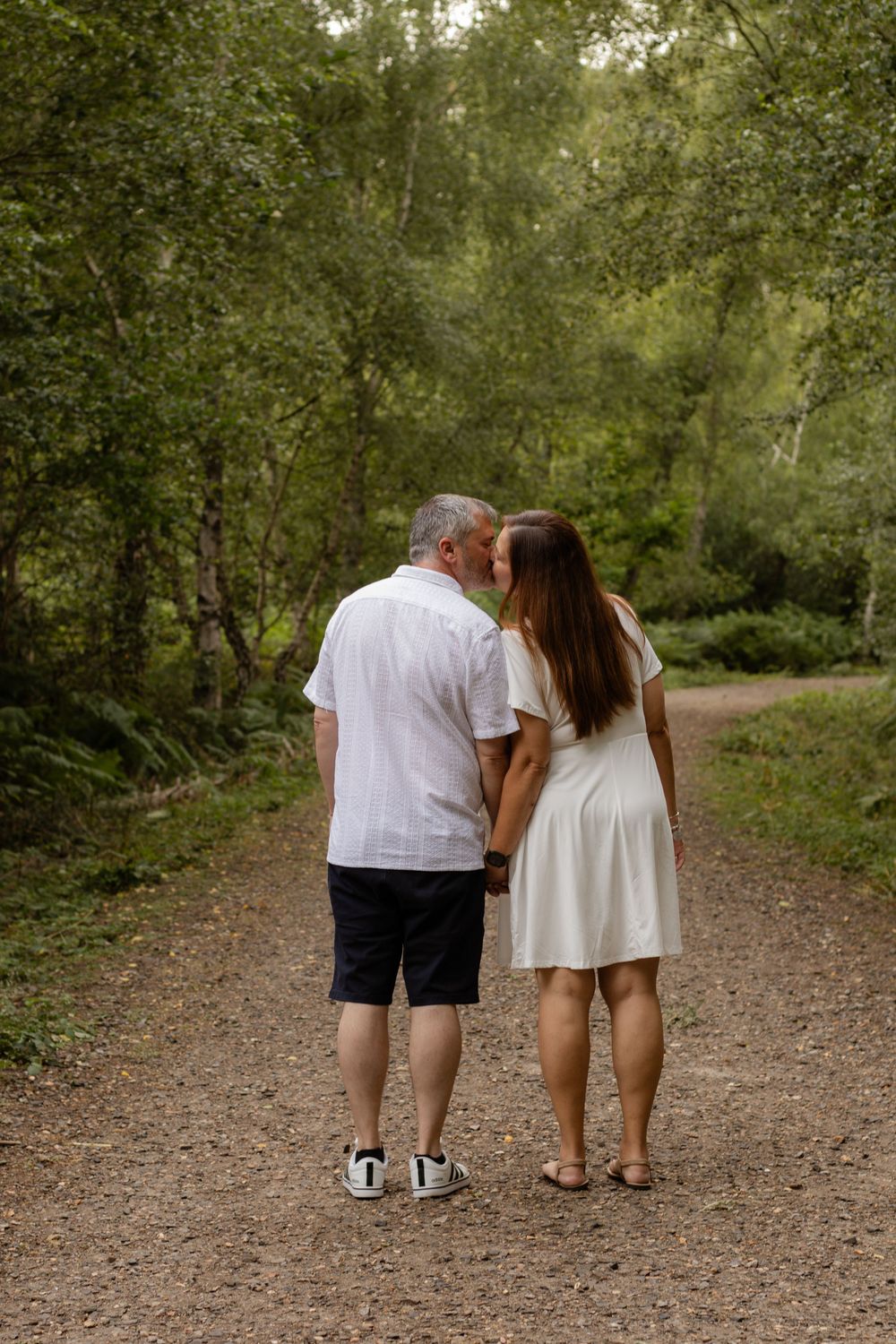 Couple walking hand in hand down a lush green forest path, sharing a kiss.