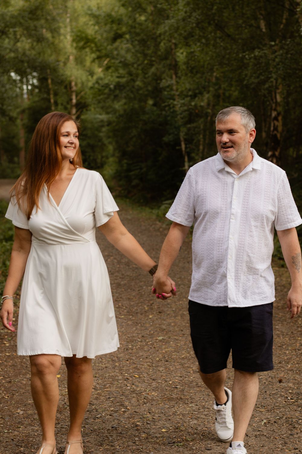 A smiling couple holds hands while walking on a forest path, woman in white dress and man in white shirt and dark shorts.
