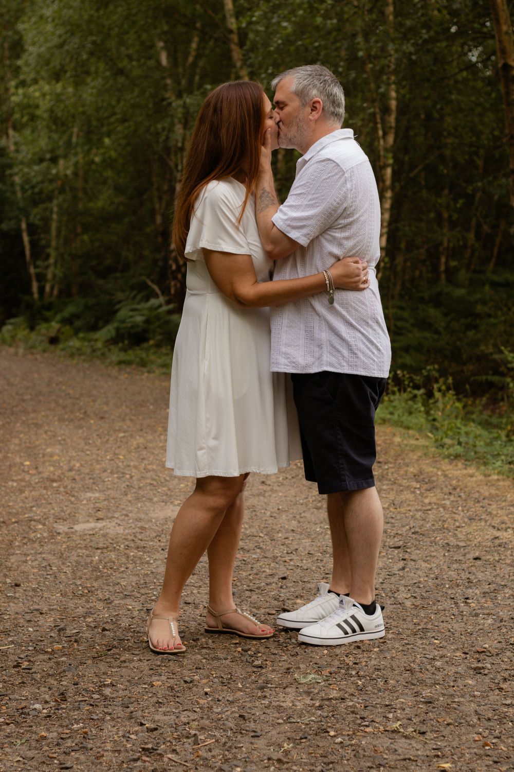 A couple kissing on a forest path, woman in white dress and sandals, man in white shirt and black shorts with sneakers.